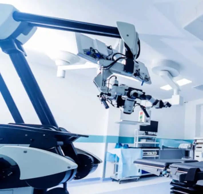 A modern surgical robot with multiple articulated arms is positioned over an operating table in a bright, sterile hospital operating room.