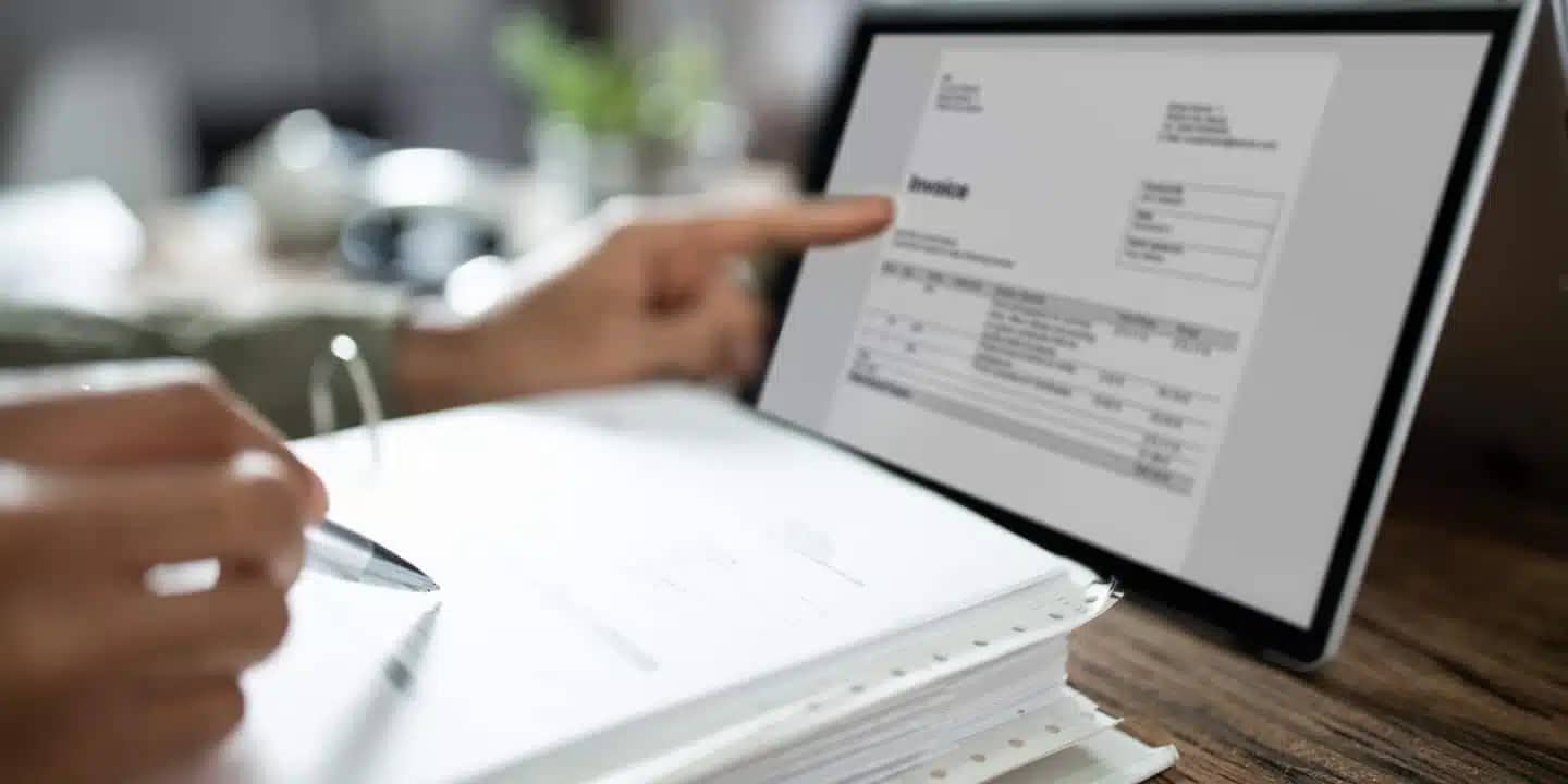 A person reviews a paper document and points at an invoice displayed on a tablet screen at a desk.