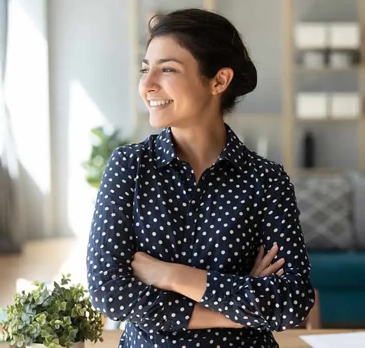 Woman with dark hair in a navy blue polka dot shirt stands indoors with arms crossed, smiling and looking to the side. A potted plant is on the table in front of her.