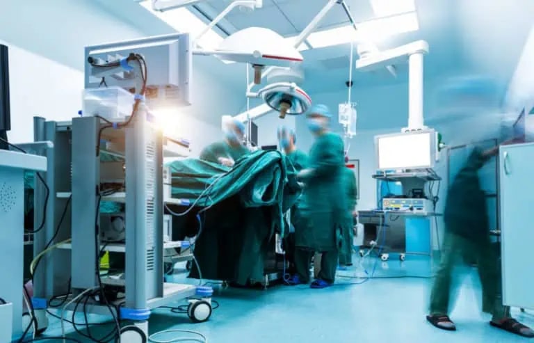 Medical team in surgical gowns performing an operation in a brightly lit operating room with medical equipment and monitors.