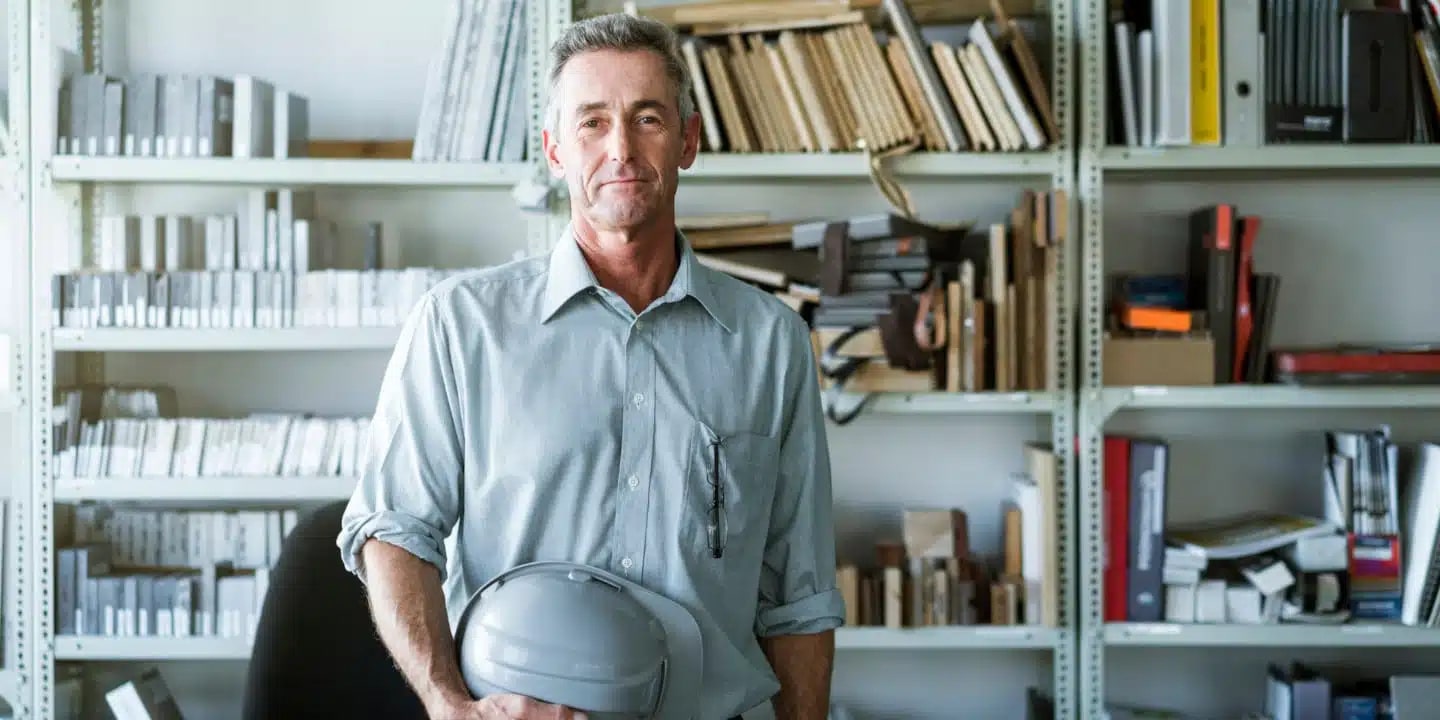 A man wearing a light blue shirt stands in front of shelves filled with books and files, holding a gray hard hat.