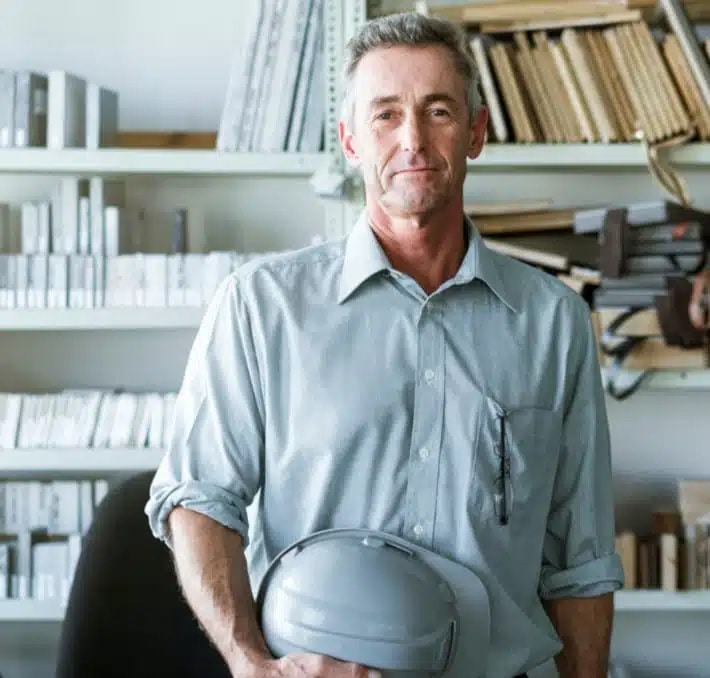 A middle-aged man in a light gray shirt stands in an office, holding a gray hard hat, with shelves of books and documents in the background.