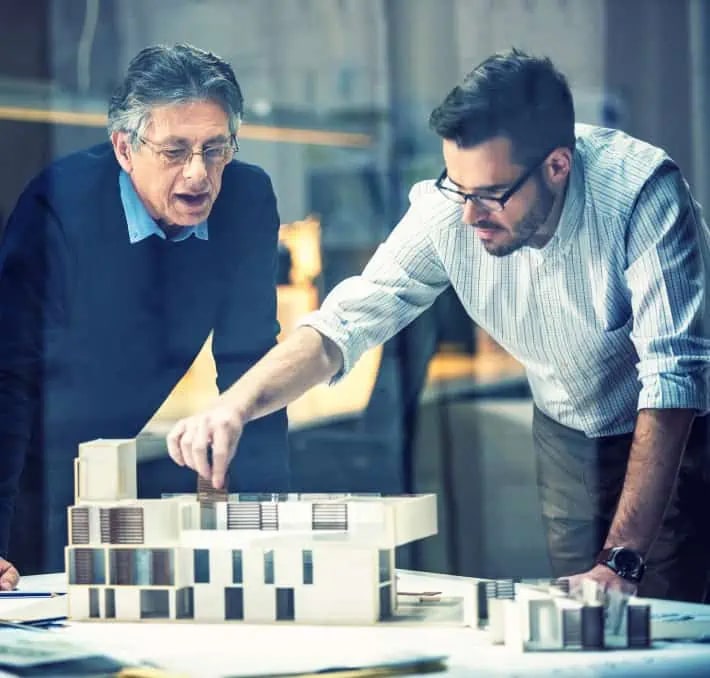 Two men examine and discuss a modern architectural model on a table, appearing focused and engaged in a professional setting.