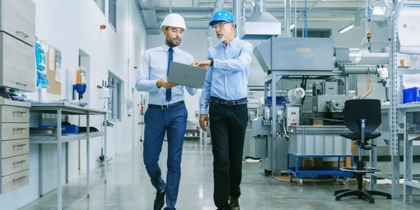 Two men in hard hats and business attire walk through a modern factory, reviewing information on a laptop. Industrial equipment and machinery are visible in the background.