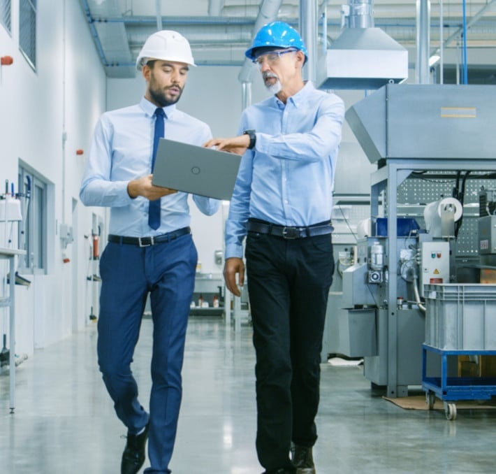 Two men wearing hard hats and business attire walk through an industrial facility while looking at a laptop.