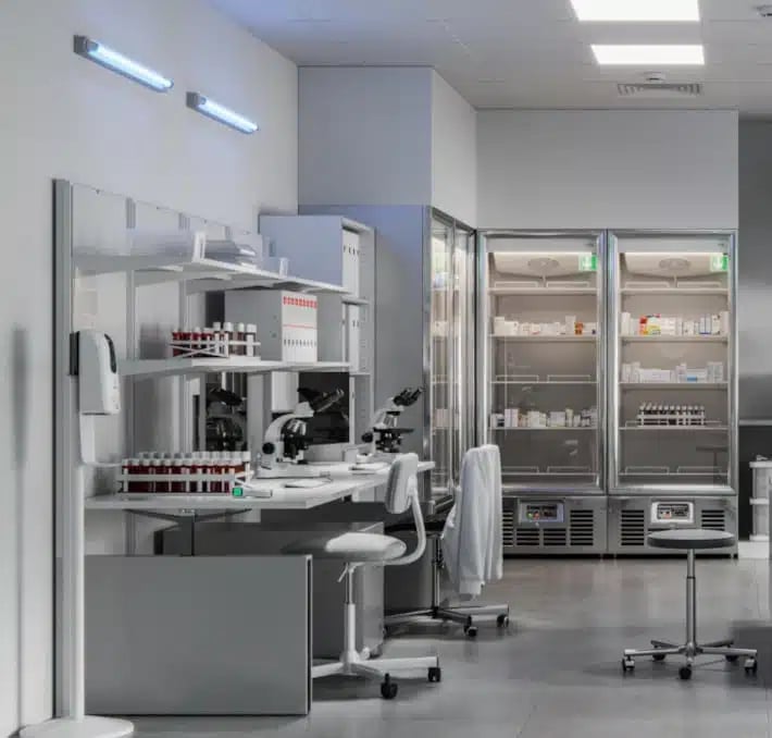 A modern laboratory with microscopes, test tubes, a white lab coat on a chair, and refrigeration units stocked with medical supplies.