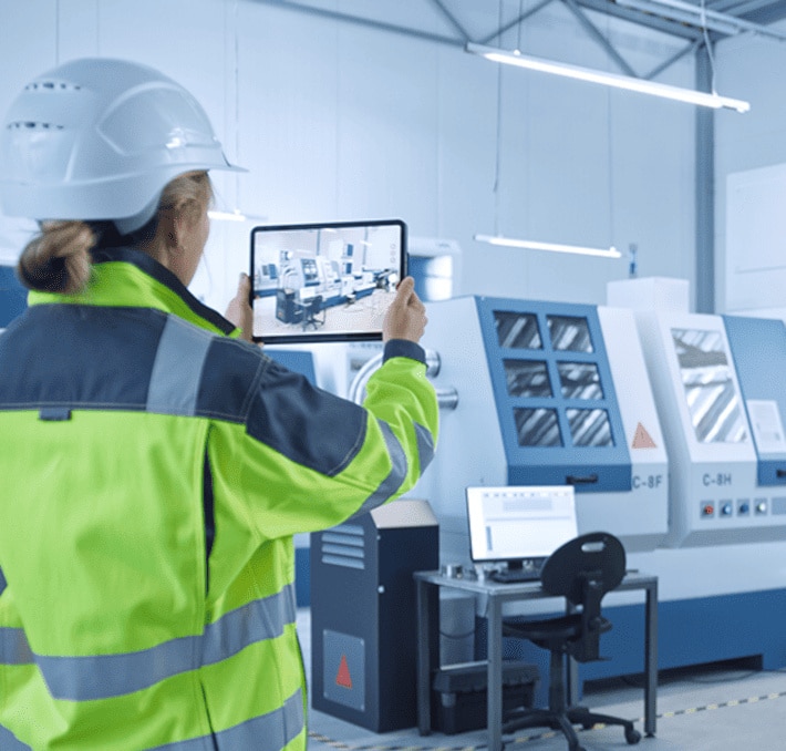 Person in safety gear uses a tablet to inspect or photograph industrial machinery inside a manufacturing facility.