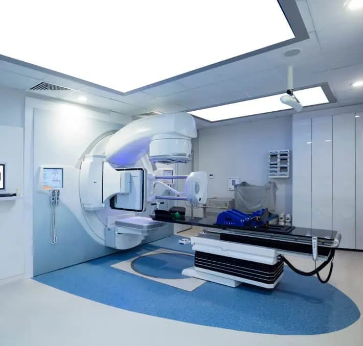 A modern hospital room with a large medical imaging or radiation therapy machine, an examination bed, and various control panels and equipment.