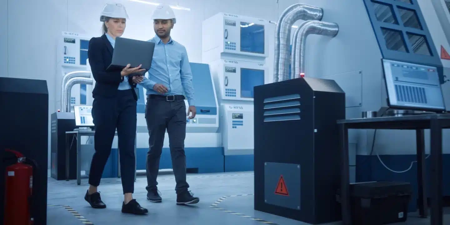 Two engineers wearing safety helmets examine a laptop while walking through a modern industrial facility with various machines and equipment visible in the background.