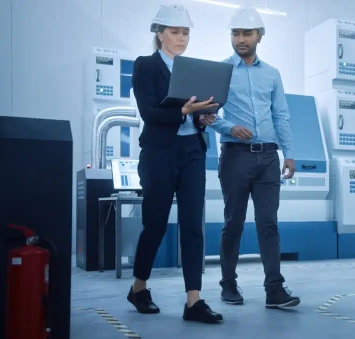 Two engineers wearing hard hats review information on a laptop while walking through a modern industrial facility with machinery in the background.