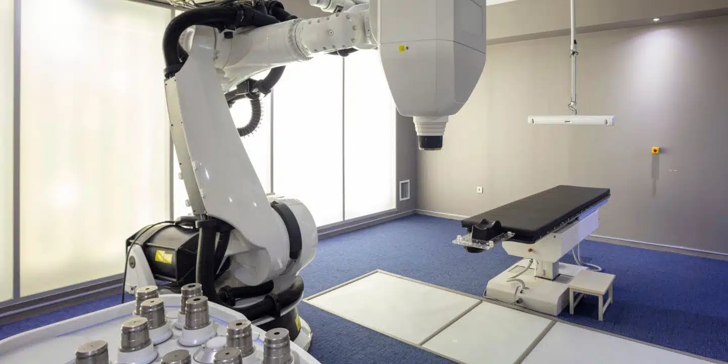 A medical robotic arm is positioned next to an empty treatment table in a modern, well-lit clinical room.