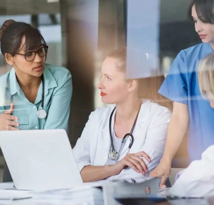Three doctors in discussion at a desk with a laptop, papers, and medical charts; two wear white coats, one wears blue scrubs.