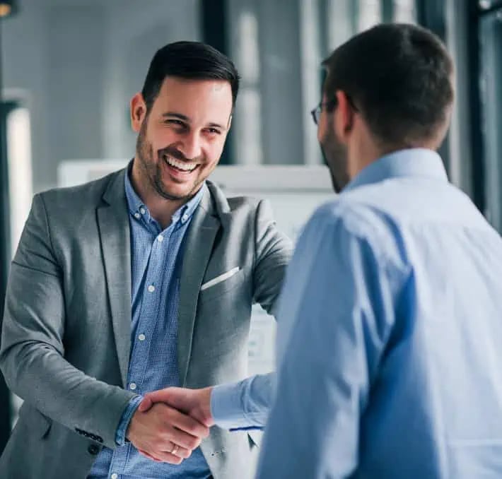 Two men in business attire shake hands and smile at each other in a modern office setting.
