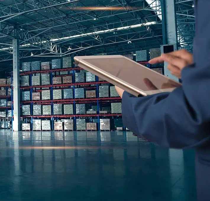 Person using a tablet in a large warehouse with stacked pallets on shelving units in the background.
