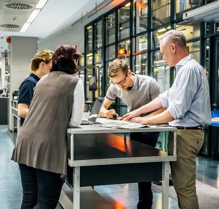 Four people stand around a table in a modern office or lab, reviewing documents and working together, with computers and equipment visible in the background.