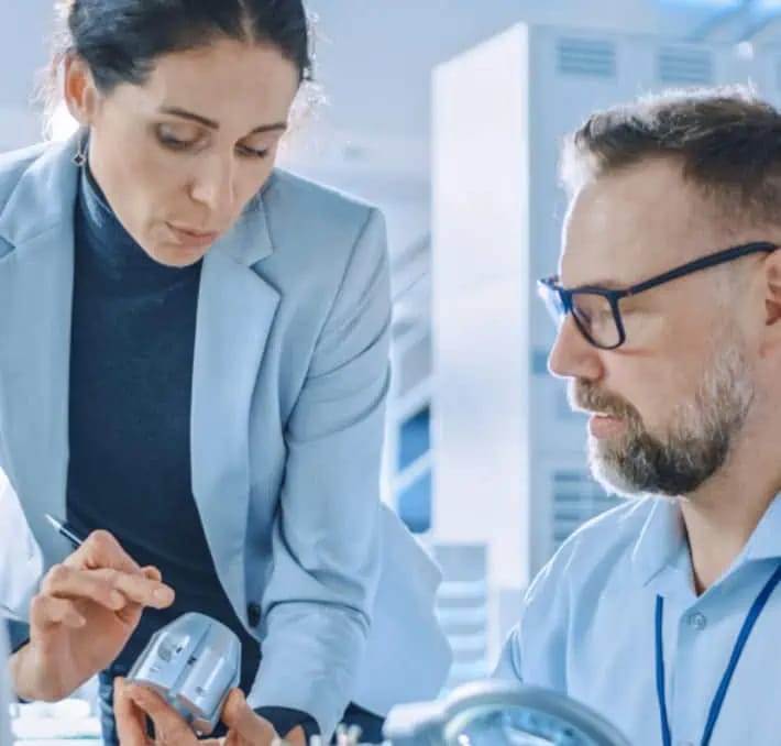 Two professionals examine a small electronic device in an office or laboratory setting, both wearing business attire and appearing focused on the task.