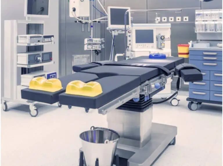 A modern operating room with an adjustable surgical table, medical equipment, monitors, storage drawers, and overhead lights.