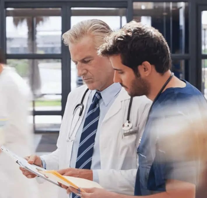 Two healthcare professionals, one in a white coat and one in scrubs, review documents together in a hospital setting.